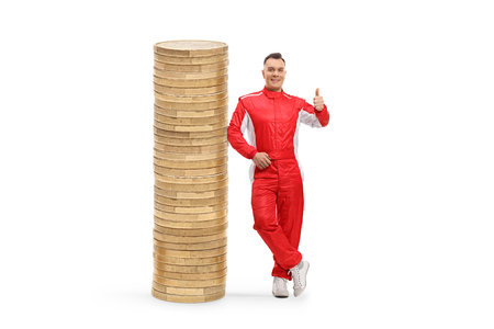 Full length portrait of a car racer in a red suit standing next to a stack of coins and gesturing thumbs up isolated on white backgroundの写真素材