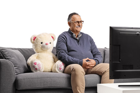 Mature man with a teddy bear sitting on a sofa and watching tv isolated on white backgroundの写真素材