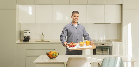 Young man in pajamas holding a breakfast tray in a kitchenの写真素材