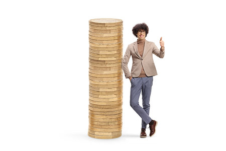 Young man gesturing thumbs up and leaning on a pile of coins isolated on white backgroundの写真素材