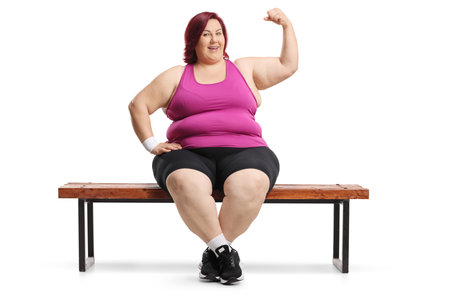 Plus size woman in sportswear sitting on a bench and showing arm muscles isolated on white backgroundの写真素材