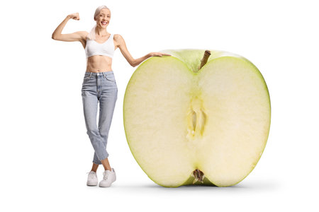 Happy slim woman in jeans and crop top standing next to a big green apple isolated on white backgroundの写真素材