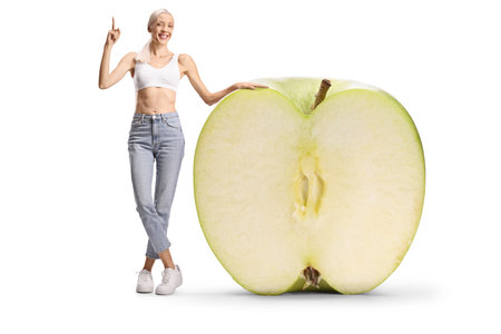 Happy slim woman standing next to a big green apple and pointing up isolated on white backgroundの写真素材