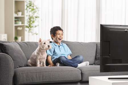 Kid and dog sitting on a sofa at home watching tv and laughingの写真素材