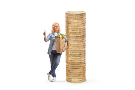 Woman with a grocery bag standing next to a pile of coins and gesturing thumbs up isolated on white backgroundの写真素材