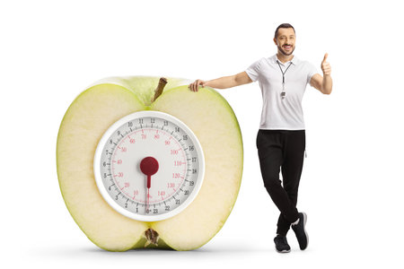 Sports coach gesturing thumbs and leaning on a weight scale isolated on white backgroundの写真素材