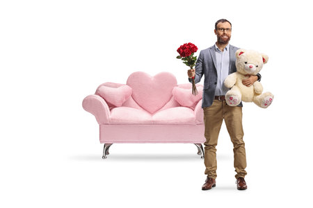 Full length portrait of a man holding a teddy bear and red roses in front of pink sofa isolated on white backgroundの写真素材