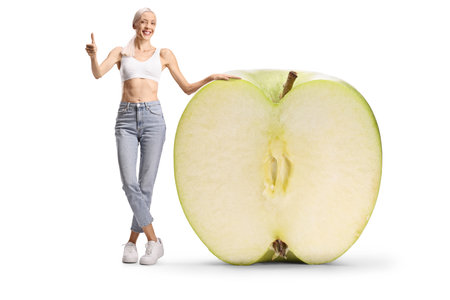 Happy slim woman in jeans and crop top standing with a big green apple isolated on white backgroundの写真素材