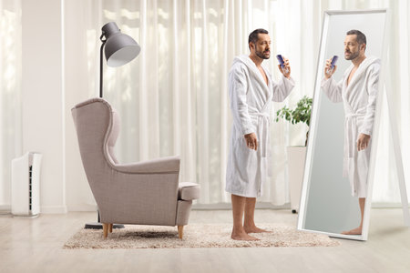 Young man in a bathrobe spraying a perfume in front of a mirror at homeの写真素材