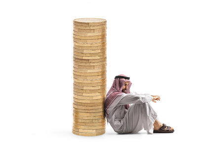 Pensive middle eastern man sitting and leaning on a pile of coins isolated on white backgroundの写真素材