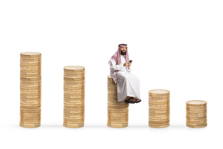 Middle eastern man using a smartphone and sitting on a pile of coins isolated on white backgroundの写真素材