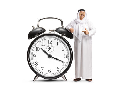Middle eastern man in traditional clothes leaning on a big alarm clock and pointing isolated on white backgroundの写真素材