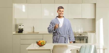 Young man in a bathrobe holding a cup in a kitchenの写真素材