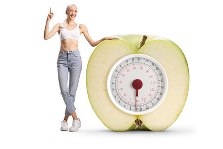 Happy slim woman in jeans and crop top pointing next to an apple weight scale isolated on white backgroundの写真素材