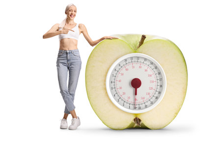Happy slim woman in jeans and crop top pointing at an apple weight scale isolated on white backgroundの写真素材