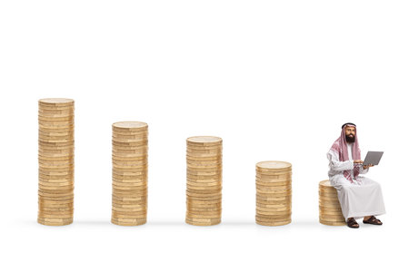 Middle eastern man with a laptop computer sitting on a pile of coins isolated on white backgroundの写真素材
