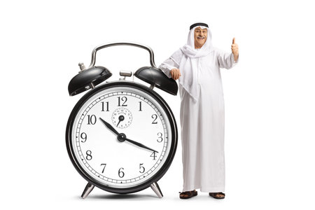 Middle eastern man gesturing thumbs up and leaning on a big alarm clock isolated on white backgroundの写真素材