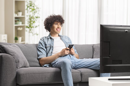 Young man sitting on a sofa in front of tv and holding a smartphoneの写真素材