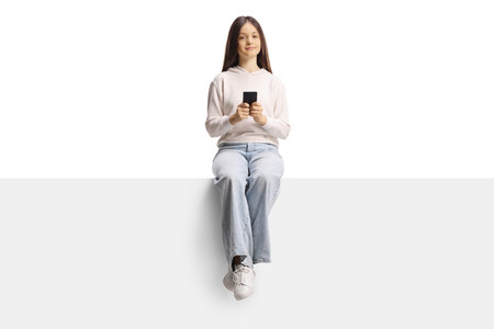Female teenage girl sitting on a blank panel and using a smartphone isolated on white backgroundの写真素材