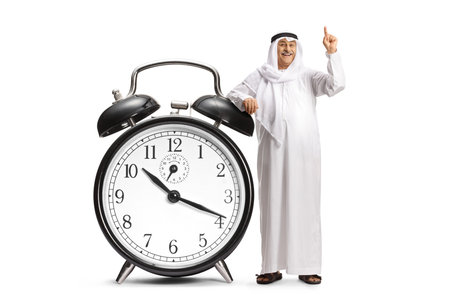 Middle eastern man pointing up and leaning on a big alarm clock isolated on white backgroundの写真素材