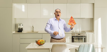 Shocked mature man in a tight shirt holding a slice of pizza in a kitchenの写真素材