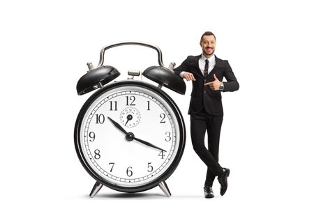 Full length portrait of a businessman pointing at a big alarm clock isolated on white backgroundの写真素材