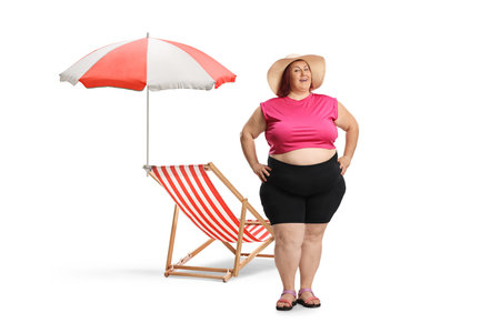 Corpulent woman in summer clothes in front of a beach chair and umbrella isolated on white backgroundの写真素材