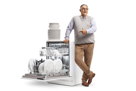 Smiling mature man leaning and pointing at a dishwasher with a pile of plates isolated on white backgroundの写真素材