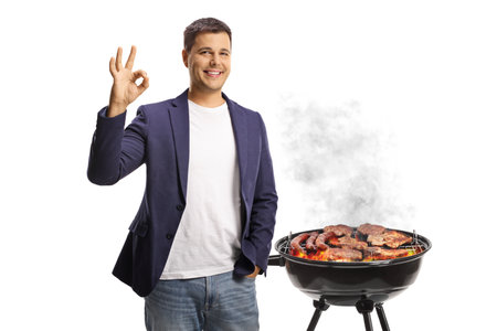 Young man gesturing ok sign in front of a bbq isolated on white backgroundの写真素材