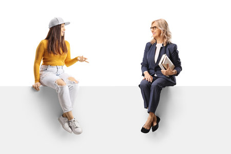 Young female sitting on a blank panel and talking to a middle aged woman isolated on white backgroundの写真素材