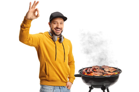 Young man with headphones gesturing ok sign in front of a bbq isolated on white backgroundの写真素材