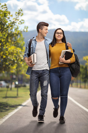 Full length portrait of two teenage students with backpacks and books walking in a parkの写真素材