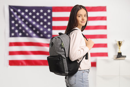 Female teenage student with a backpack and book standing in front of a USA flag
