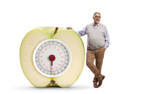 Full length portrait of a corpulent mature man leaning on an apple with a weight scale isolated on white backgroundの写真素材