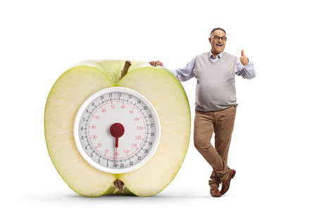 Corpulent mature man leaning on an apple and weight scale and gesturing thumbs up isolated on white backgroundの写真素材