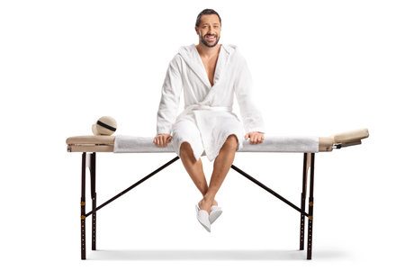 Man in a white bathrobe sitting on a massage table isolated on white backgroundの写真素材