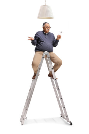 Confused mature man holding a light bulb and sitting on a ladder under lampshade isolated on white backgroundの写真素材