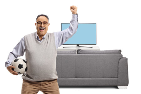 Elderly football fan cheering with a ball in front of tv and sofa isolated on white backgroundの写真素材