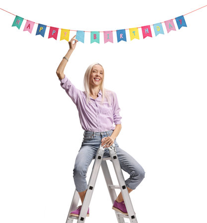 Woman sitting on a ladder and hanging birthday decorative flags isolated on white backgroundの写真素材