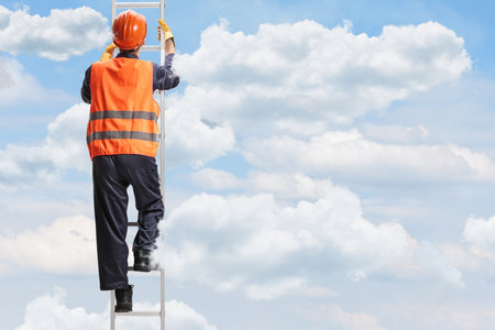 Rear view shot of a worker in a reflective vest and helmet climbing a ladder in the skyの写真素材