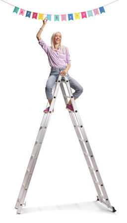 Woman sitting on top of a ladder nd hanging birthday decoration flags on ceiling isolated on white backgroundの写真素材