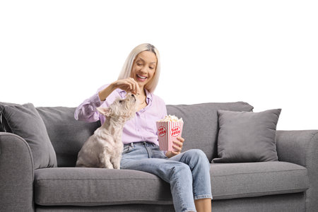 Woman sitting on a sofa and giving popcorn to a dog isolated on white backgroundの写真素材