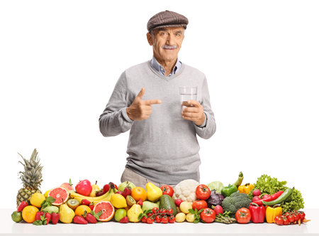 Male pensioner with a pile of fruits and vegetables pointing at a glass of water isolated on white backgroundの写真素材