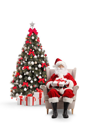 Santa claus sitting in armchair with a present in front of a Christmas tree isolated on white backgroundの写真素材