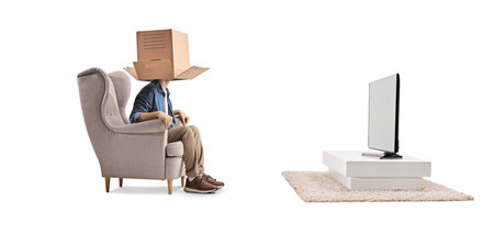 Man sitting in an armchair in front of tv with a cardboard box over head isolated on white backgroundの写真素材