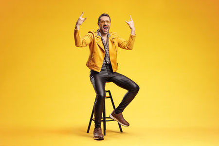 Excited man doing a rock hand sign seated on a stool against yellow background