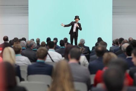 Performer in a black tuxedo and top hat speaking to a large audience in a conference hall