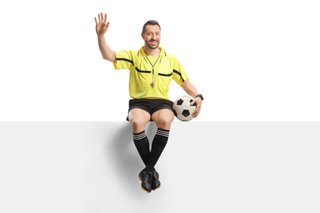 Football referee sitting on a blank panel with a ball and waving isolated on white backgroundの写真素材