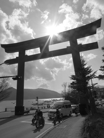 Vehicles line up and drive under a large Japanese shinto gate with the sun setting and nature in the background.のeditorial素材