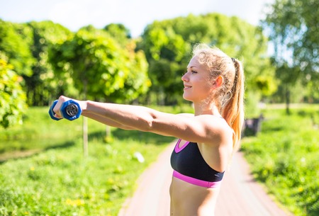 Young woman training with dumbbells in city park at summer day.